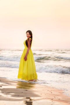 A girl in a long yellow dress walks by the sea in the early morning at dawn.