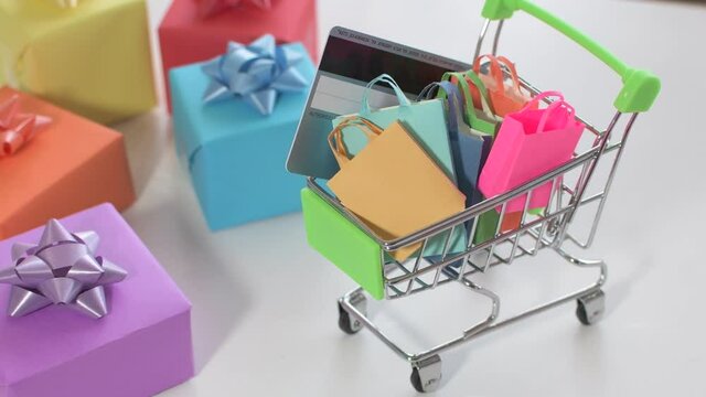 Supermarket trolley with shopping bags and credit card, next to small colored gifts, on white background.