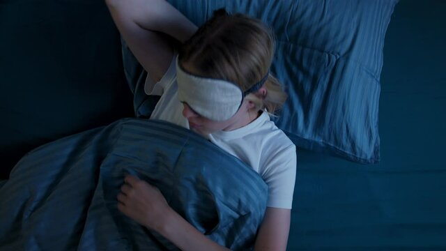 Young woman in bed in sleep mask, suffers from insomnia, tries to fall asleep, tosses, removes sleep mask from eyes and discontentedly look camera