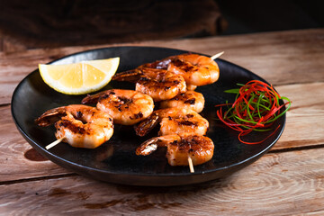 Appetizing fried shrimp on a skewer in a black plate on a wooden surface among lemon and seasonings. The concept of fast and wholesome food