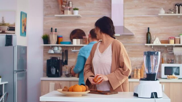 Couple making smoothie in kitchen. Woman peeling of orange while smiling at husband. Cheerful family making together organic healthy fresh nutritious tasty juice for breakfast from fresh fruits while