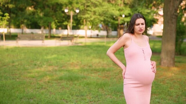 A pregnant girl is standing in the park, and her stomach begins to hurt. a pregnant woman strokes her stomach and walks from side to side