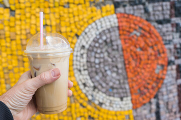 Holding iced coffee in takeaway cup on . wall background with colored mosaics