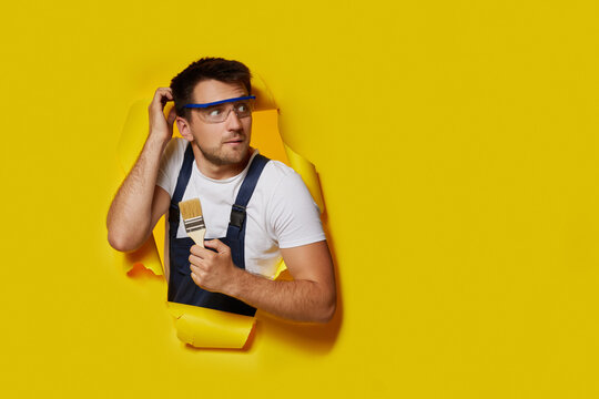 caucasian painter worker in safety glasses and uniform holding paint brush through hole in yellow paper. copy space. handyman thinking an idea. Skeptic and doubts concept.