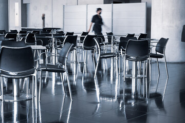 Modern chairs and tables stand in a cool urban space on a dark reflective background and a man with a face mask walks in the background.