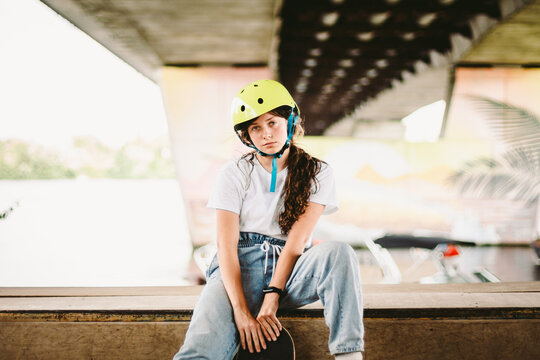 Teenage girl in helmet and stylish clothes posing on half pipe ramp an outdoor skate park. Beautiful kid female model skateboarder with skate board in urban extreme park. Schoolgirl after school