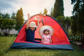 Two happy little kids boy and girl brother and sister are playing in a red camping tent in the home yard on the grass.