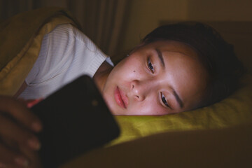 Young Asian woman using smartphone mobile surfing internet and social media on bed in bedroom at night