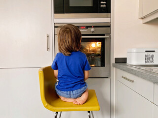 Little hungry toddler boy sitting at oven looking inside through the glass waiting for pizza.