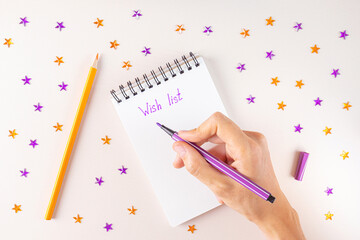 Spiral notebook and female hand with purple marker. On white sheet of paper inscription - Wish List. Around - purple and orange rhinestones in shape of stars, pencil. Top view, flat lay, copy space.