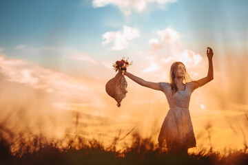 Ginger girl standing in the field with her arms up in the air, holding a hat.