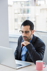 Fototapeta na wymiar Manager uomo moro vestito con camicia blu e giacca nera porta degli occhiali da vista mentre lavora seduto nella sua postazione del suo ufficio 