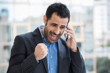 Manager uomo moro vestito con camicia blu e giacca nera parla al telefono con aria infuriata  in piedi vicino a una finestra nel suo ufficio 