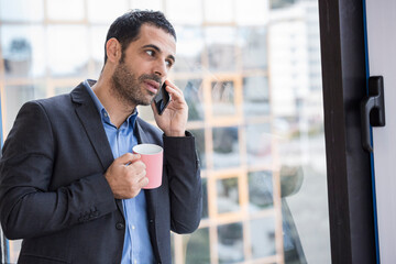 Manager uomo moro vestito con camicia blu e giacca nera parla al telefono in piedi vicino a una...