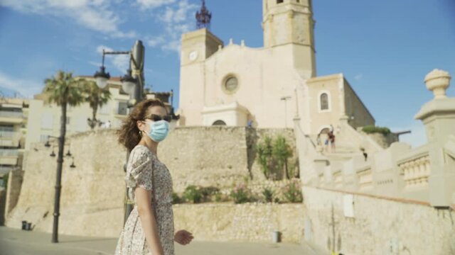 Beautiful curly brunette tourist with a surgical mask during Covid-19 in front of the Church of of Sant Bartomeu and Santa Tecla in Sitges. Safe Travel in the new normal concept