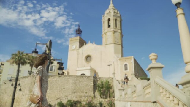 Beautiful curly brunette tourist with a surgical mask during Covid-19 in front of the Church of of Sant Bartomeu and Santa Tecla in Sitges. Safe Travel in the new normal concept