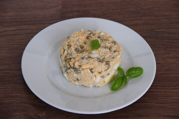 Homemade salad with cod liver, egg and cheese, dressed with mayonnaise and decorated with Basil, on a white plate and a wooden background