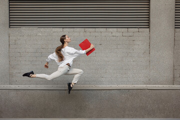 Paperwork. Jumping young woman in front of city building wall, on the run in jump high. Hurrying up to daily routine inspired and sportive. Young ballet dancer in casual clothes and sunshine.