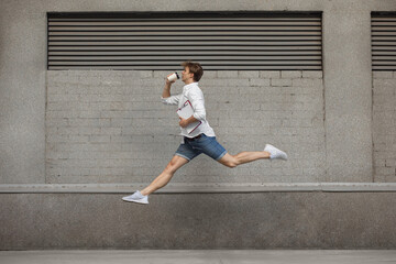 Coffee drinking. Jumping young man in front of city building wall, on the run in jump high. Hurrying up, moving to daily routine inspired and sportive. Young ballet dancer in casual clothes and