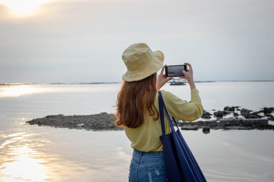 Woman standing behind and using a handheld camera to capture the sunset on the beach at sea The sea along the coast where fishing boats survive on outdoor holidays.