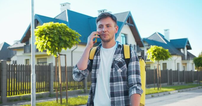 Handsome Caucasian courier with a backpack delivering the order. Smiling delivery man walking down the street calling and talking to the customer to fing the address. Delivery service during pandemic.