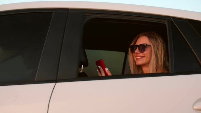 A beautiful girl with a phone in her hands rides in a car and looks out the window. Beautiful young girl in sunglasses rides in a car holding a phone, view from another car