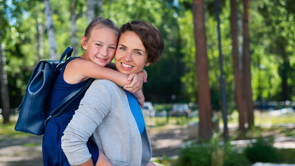 Naklejka premium A beautiful caucasian woman stands outdoors and holds daughter on her back. A cheerful smiling girl sits on her mother back while walking in the park. Friendly loving family.