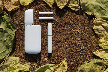 White battery  with  individual stick e-cigarette and cigarette for it  lies on  dry  tobacco texture with green, yellow  leaves  of tobacco.