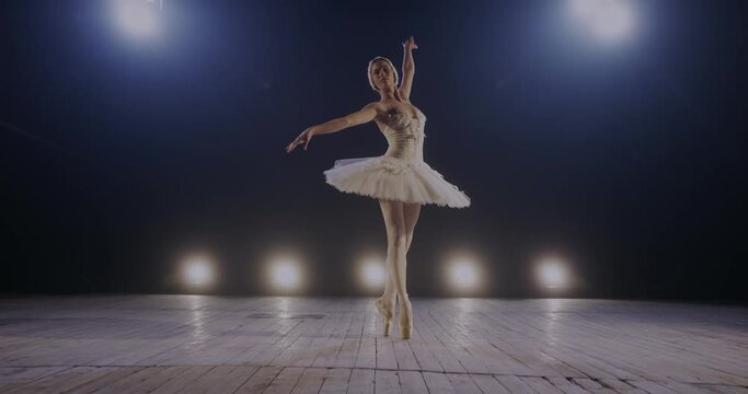 Graceful young female ballerina in tutu dancing charmingly and doing pas in light of blue light projectors on stage of theater. Camera zooming in woman dancer of ballet in lamps lights.