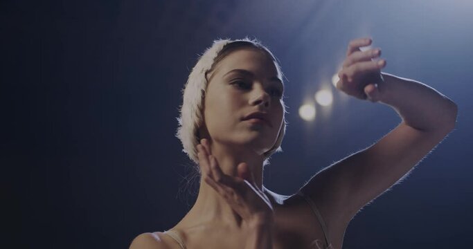 Portrait of young pretty Caucasian ballerina in crown rehearsing hands pas in darkness and ligh of lamps. Close up of female ballet dancer dancing gracefully and looking at camera.