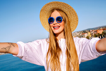 Stylish pretty blonde woman with long hairs, wearing trendy sunglasses and straw hat, making selfie in front of sea and European city, traveling summer mood.
