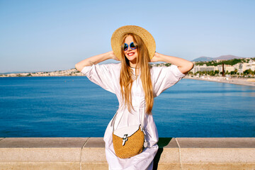 Summer lifestyle portrait of pretty blonde tourist woman posing in view point in French Riviera, wearing fashionable summer dress and luxury straw accessories, vacation in Nice.