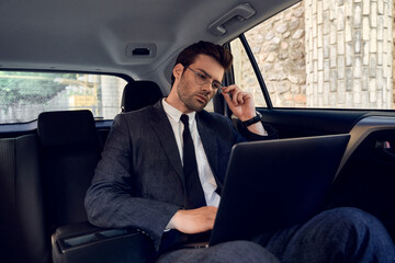 Confident young businessman seriously and thoughtfully looking at the laptop holding glasses with one hand while sitting in the car