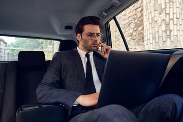 Confident young businessman seriously and thoughtfully looks at the laptop holding his chin with one hand while sitting in the car.