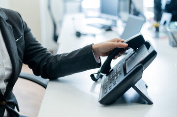 Closeup female hand on landline phone in office. Faceless woman in a suit works as a receptionist answering the phone to customer calls.