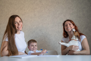 Obraz na płótnie Canvas Happy family stays at home. Two women help the boy do school homework. Lesbian couple sitting at the table with their son and a cheerful puppy. Same-sex marriage with a child. Distance learning.