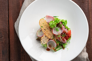 Fresh salad of greens, vegetables, radishes, cucumbers and ham in a white plate on a wooden background