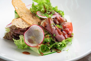 Fresh salad of greens, vegetables, radishes, cucumbers and ham in a white plate on a wooden background