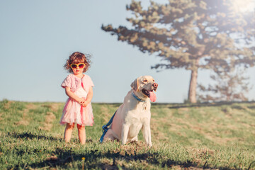 Cute stylish little Caucasian child girl in sunglasses walking dog in park field outside on summer sunny day. Kid playing with a domestic animal pet. Happy childhood concept. Chic sassy girl baby.