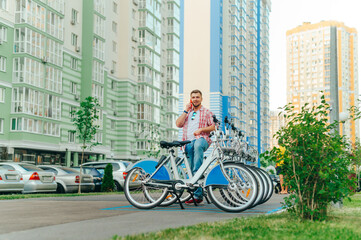 Happy young man with a beard stands near a row of sharing bicycles and talks on the phone with a smile on his face. A smiling tourist takes a bicycle for a walk