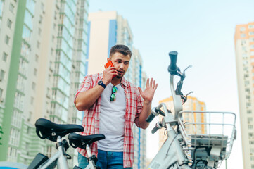 Bearded adult stands by a bicycle against the backdrop of buildings and the sky, talking on a smartphone with a serious face. Businessman speaks on the phone while cycling