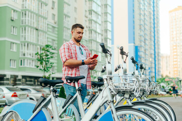 A man with a beard and a serious face uses a program on a smartphone for sharing bicycles, standing...