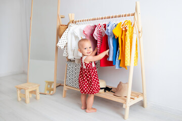 Baby girl stands near a wardrobe and chooses a dress and smiles. Dressing closet with clothes arranged on hangers. Colorful wardrobe of newborn, kids, toddlers, babies full of all clothes