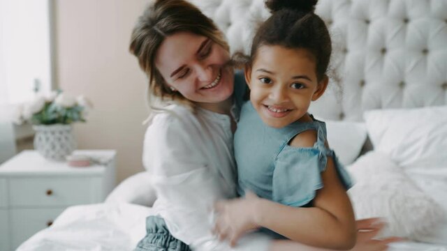 Little beautiful playful African girl daughter runs jumping to her mother nanny babysitter on large light bed in modern bedroom hugging and kissing her. Concept of friendly, happy, smiling family.