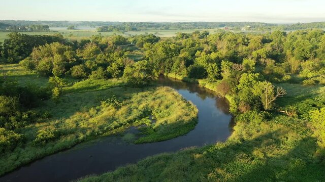 Aerial view of rural landscape with lush greens, trees, river and fog over farm fields in the background. American countryside scenery in the early sunny morning, summertime. Camera moves forward