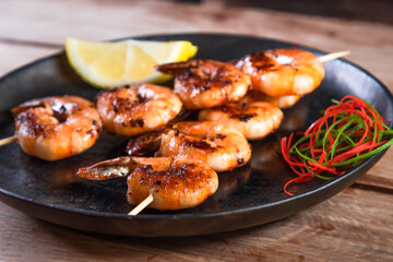 Appetizing fried shrimp on a skewer in a black plate on a wooden surface among lemon and seasonings. The concept of fast and wholesome food