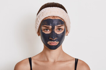 Young beautiful woman in black mud mask for face doing cosmetic procedures, looks sad, posing against white wall, wearing hair band, looks at camera.