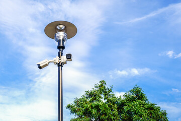 Two white surveillance cameras on the metal lamp post on blue sky background in public park.