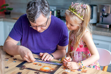 Fototapeta premium Father and daughter assemble the puzzle while sitting at the table. Games during rain and bad weather, quarantine. Family relaxing educational games at home.
