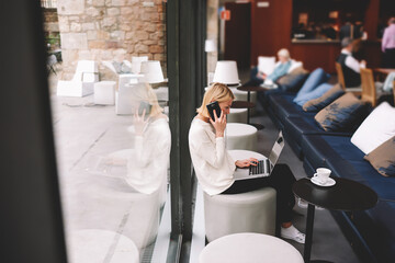 Young businesswoman sitting front open laptop computer and talk on smartphone, female person using net-book during morning breakfast in modern coffee shop, freelancer working on the distance in cafe